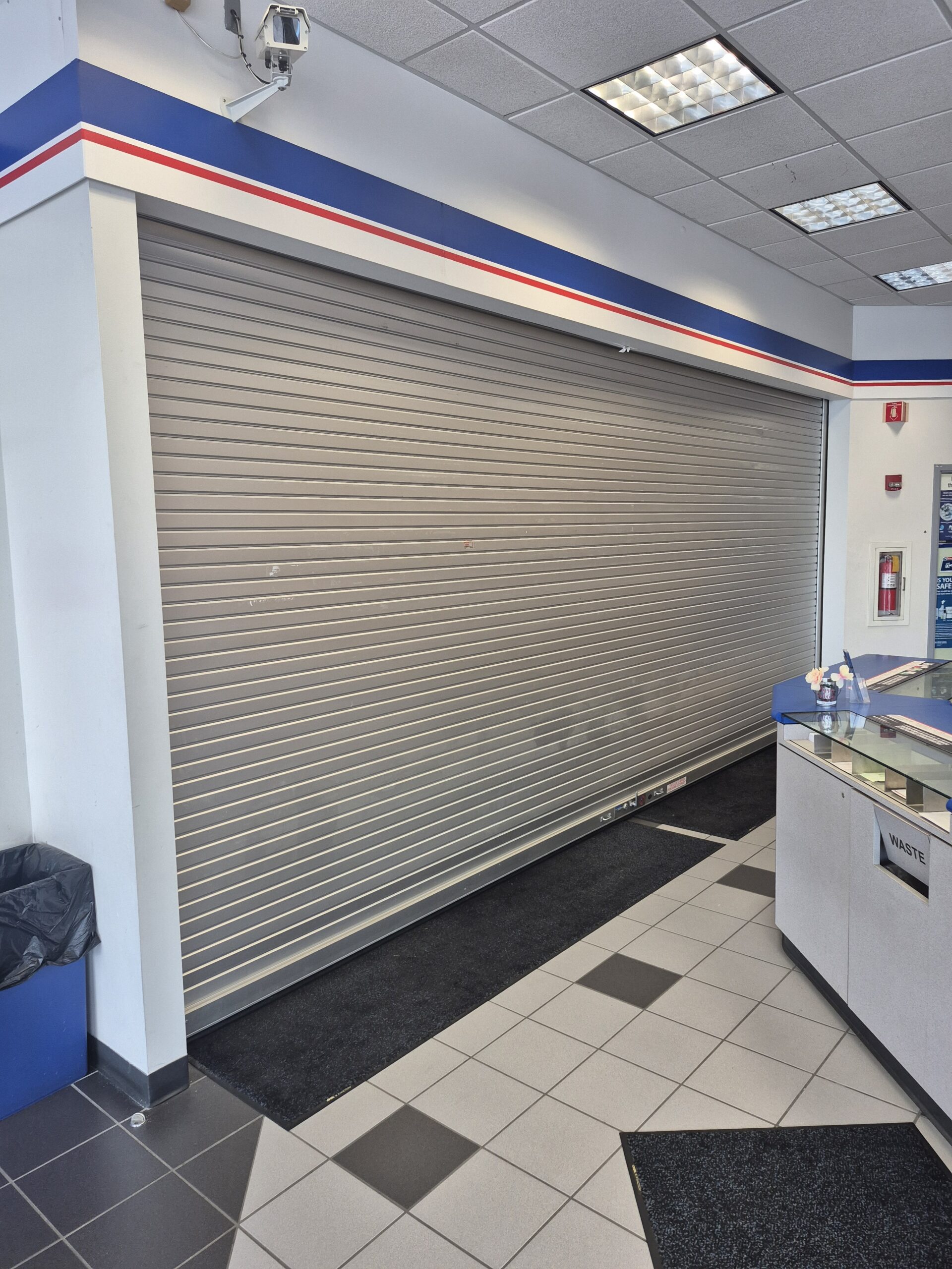 A closed metallic security shutter inside a commercial or retail space, with tiled flooring and a counter visible on the right side.