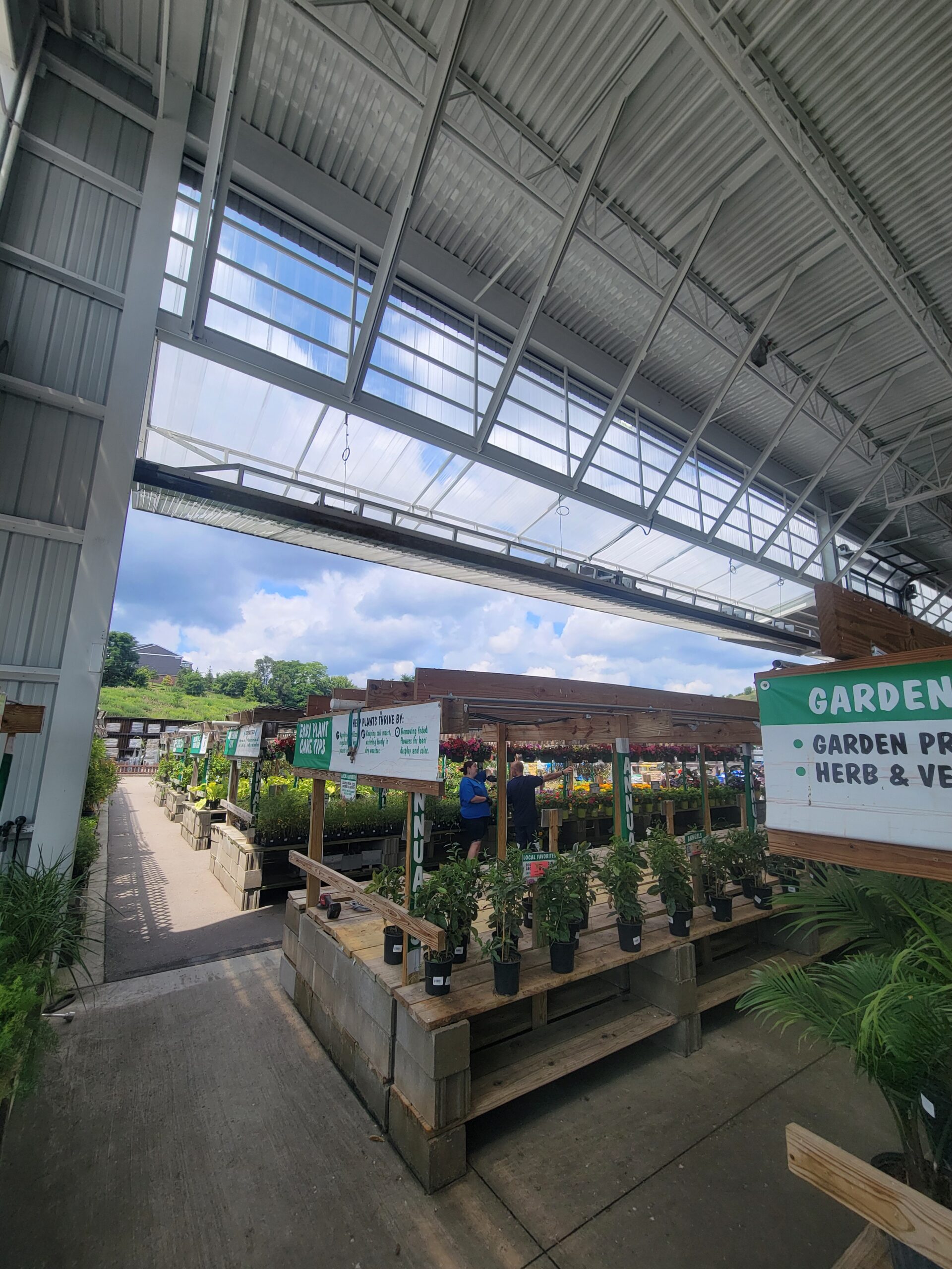 Indoor garden center with potted plants displayed on wooden tables, open roof panels, and signs indicating garden products, herbs, and vegetables.