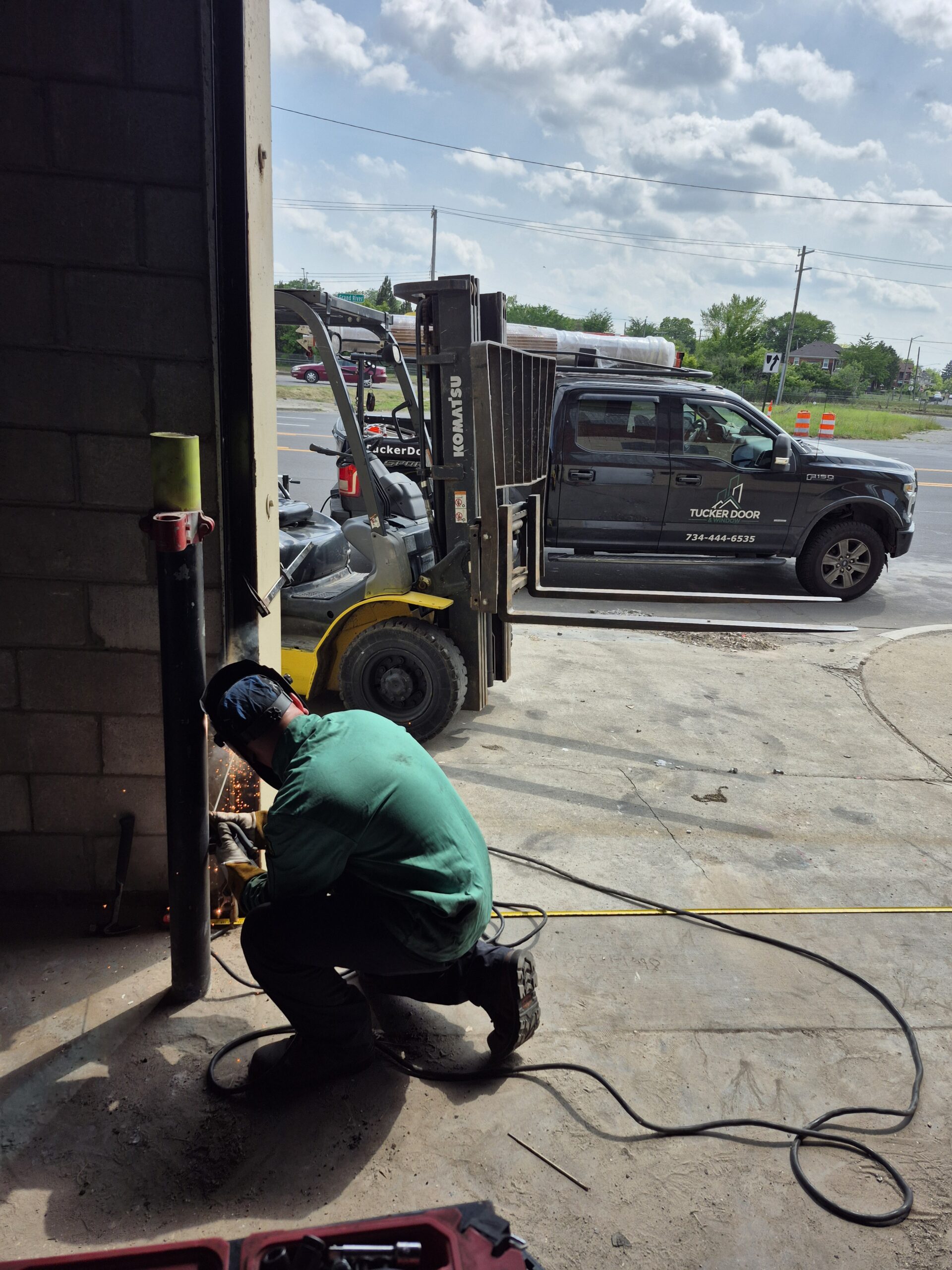 A worker in a green shirt and helmet welds a pipe near an open garage door, with a forklift and a parked truck visible outside in daylight.
