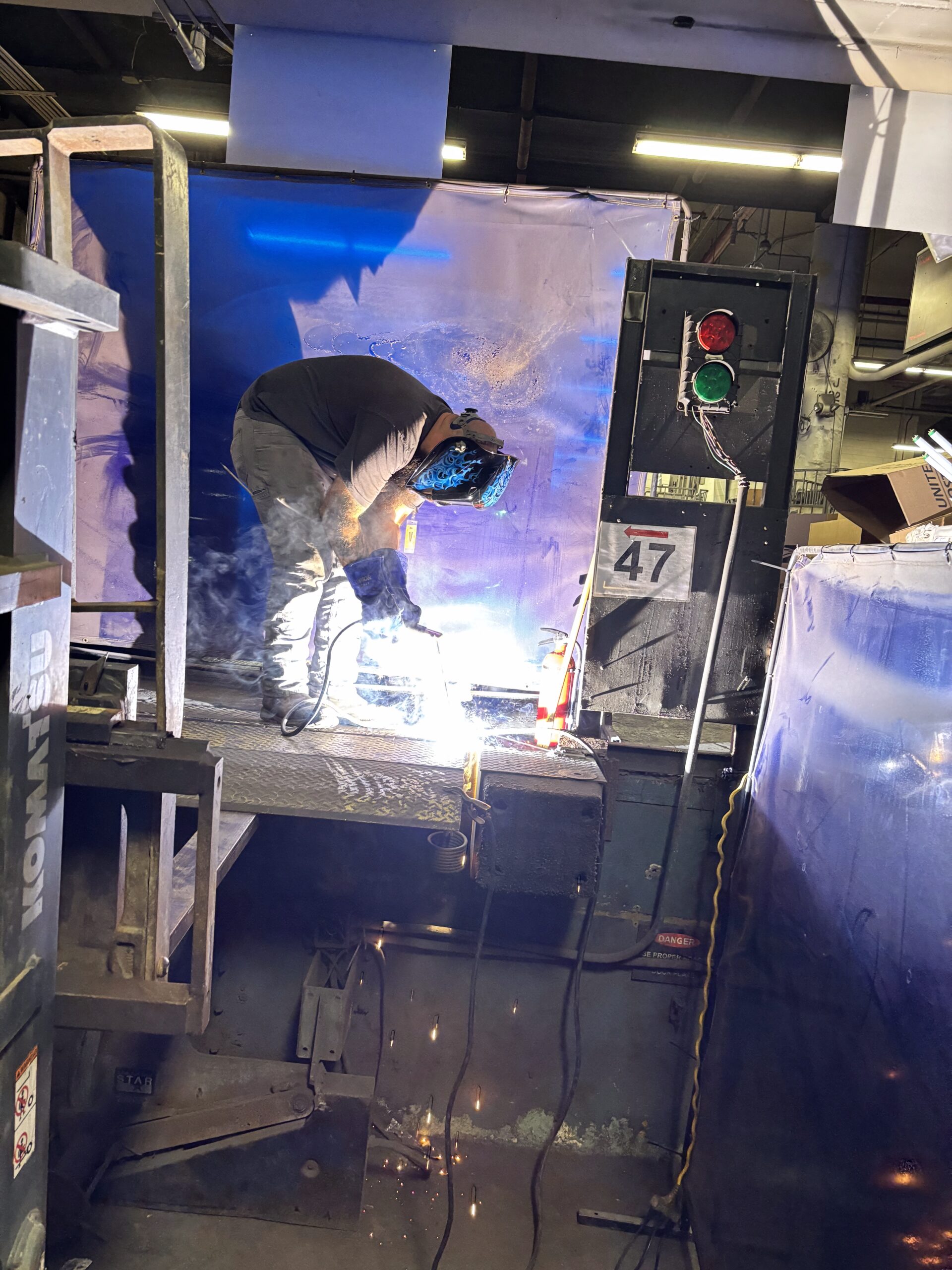 A person wearing protective gear welds metal on an elevated platform in an industrial setting, with sparks visible and a lit welding torch.