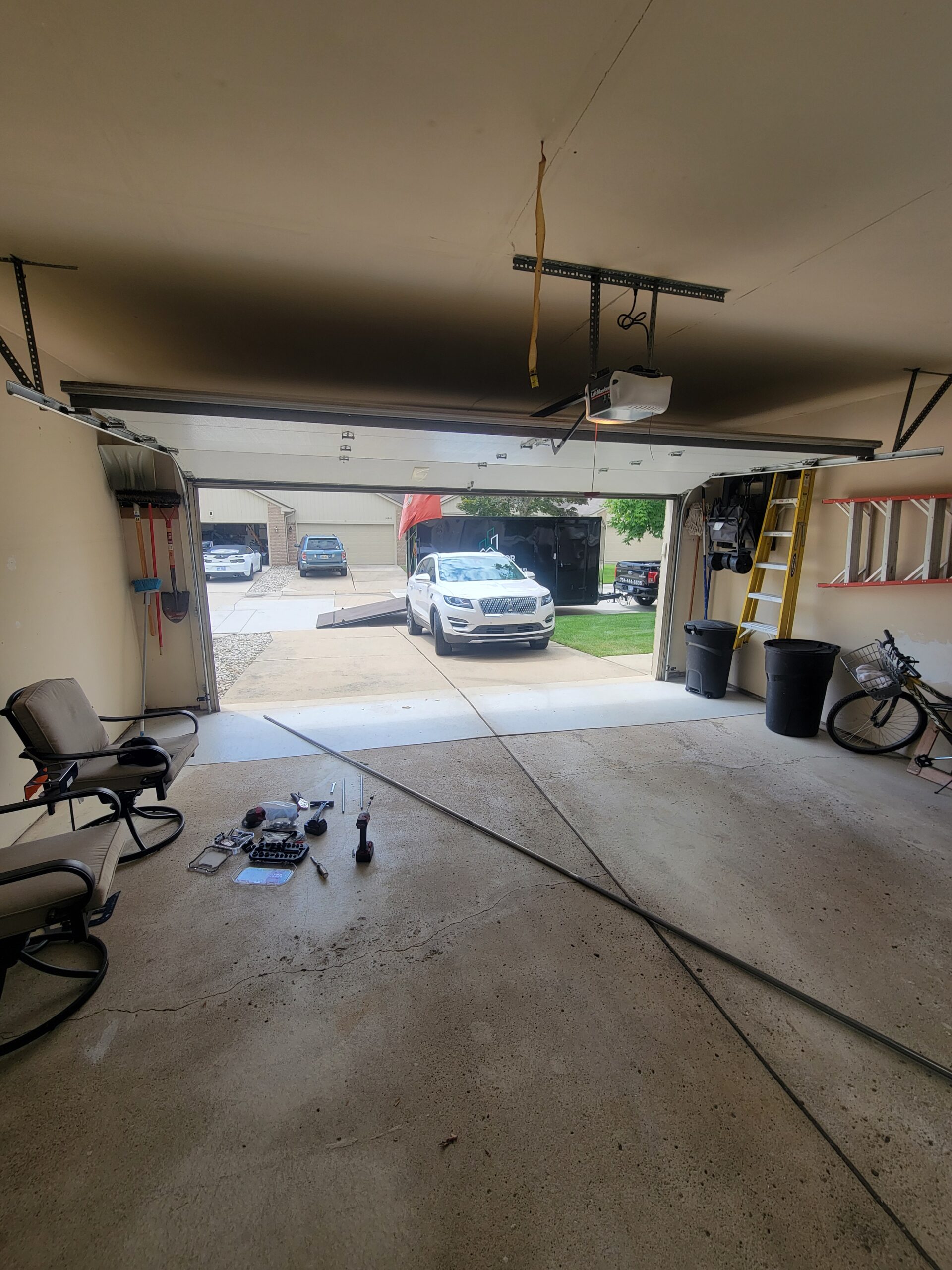 Open garage with tools and parts on the floor, a car parked on the driveway, and bicycles and chairs visible inside the garage.